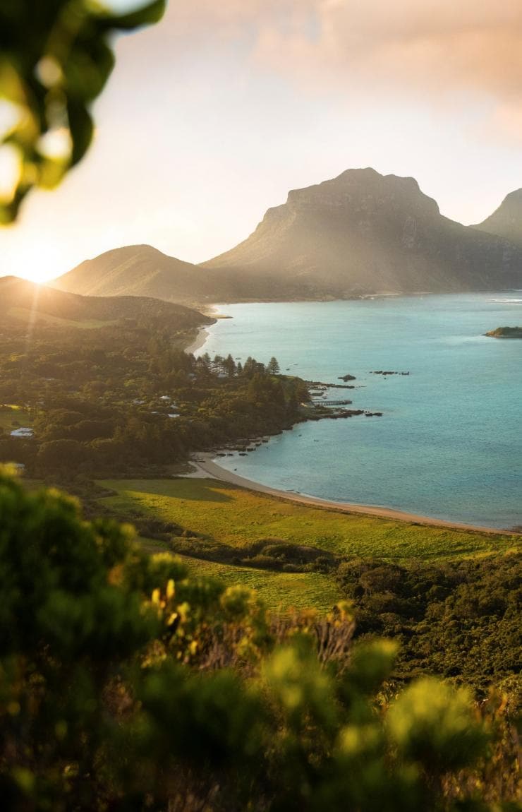 A tree-covered island with bright blue ocean and two towering mountains in the distance at Mount Lidgbird and Mount Gower, Lord Howe Island, New South Wales © Tom Archer