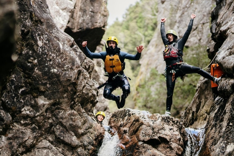 Two people wearing helments at life vests are seen at the top of a small waterfall as one of them jumps from the rock while the other cheers behind them with Cradle Mountain Canyons, Cradle Mountain, Tasmania © Tourism Australia