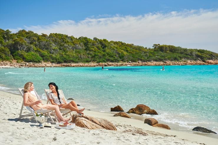 Two women lounging on beach chairs with champagne and a charcuterie board while people stand up paddle board through clear blue water in the background at Meelup Beach, Dunsborough, Western Australia © Frances Andrich