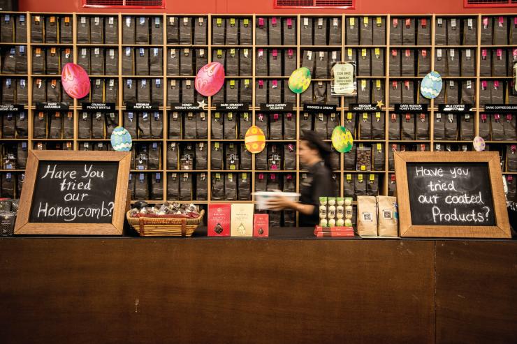 A staff member at Margaret River Chocolate Company walking past a counter and shelves filled with chocolate at Swan Valley, Western Australia © Tourism Western Australia