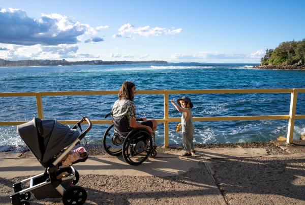 A person in a wheelchair seated with their child on a waterfront overlooking the ocean in Manly, Sydney, New South Wales © Tourism Australia
