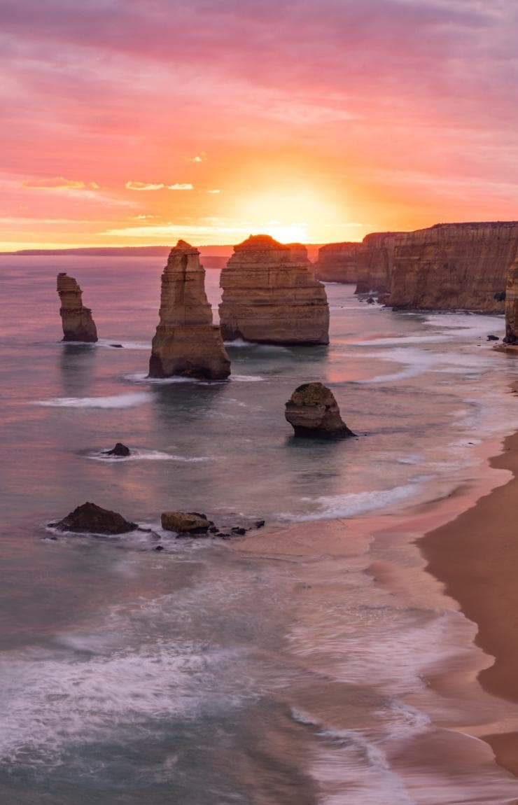 Vibrant pink and orange sunset skies illuminate towering limestone stacks along a sandy beach and steep coastal cliffs with Oceania Tours and Safaris, 12 Apostles, Great Ocean Road, Victoria. © Tourism Australia