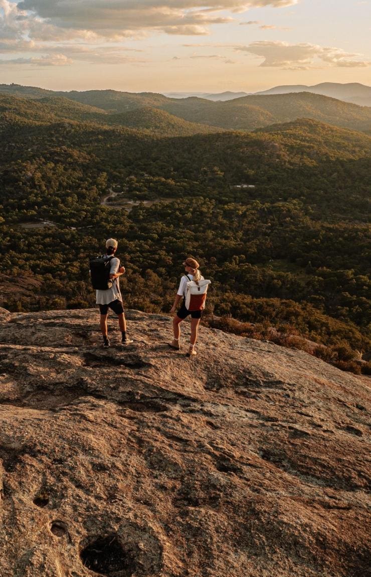 Two hikers stand atop a rocky outcrop looking over Girraween National Park, Queensland Country, Queensland © Tourism Australia