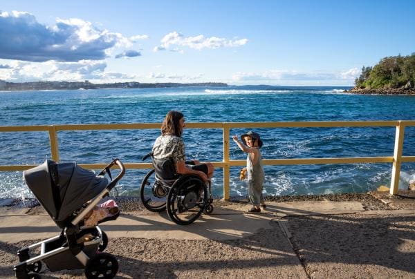 A person in a wheelchair seated with their child on a waterfront overlooking the ocean in Manly, Sydney, New South Wales © Tourism Australia
