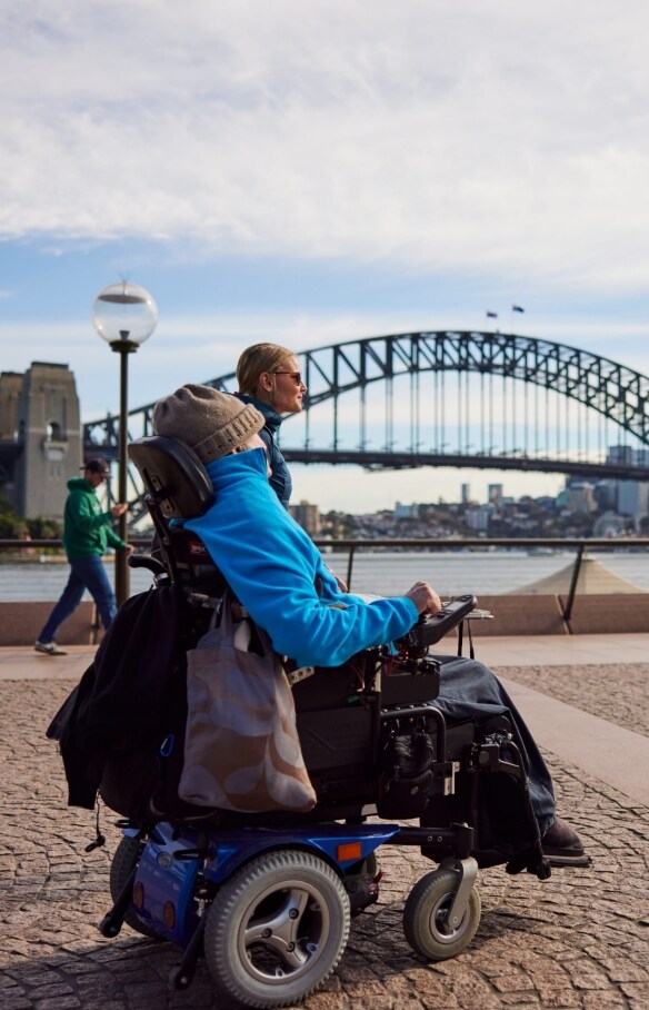 Person using a mobility device with another person walking beside them approaching the Sydney Opera House with the Sydney Harbour Bridge in the background in Sydney, New South Wales © Destination NSW