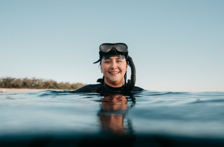 Amy Gash smiling in the ocean while wearing a snorkel with a tree-covered island in the distance, Lady Elliot Island, Southern Great Barrier Reef, Queensland © Tourism Australia