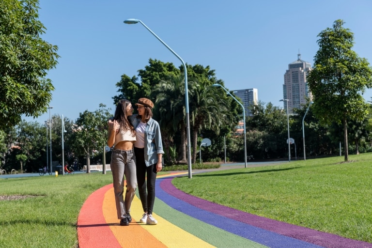 A couple with their arms around one another as they wander along a rainbow pathway through the green grass and trees of Prince Alfred Park, Surry Hills, New South Wales © Destination NSW