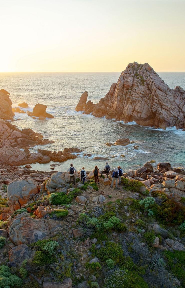 An aerial view captures a group of hikers walking along the cliff edge near Sugarloaf Rock in Margaret River. The setting sun bathes the rugged rocks and headlands in a warm, golden glow. Gentle waves lap against the coastline below as the hikers pause to take in the breathtaking scenery. Midway through their 4-day Cape to Cape Walk, they are surrounded by pristine coastal beauty under clear, expansive skies. © Tourism Australia
