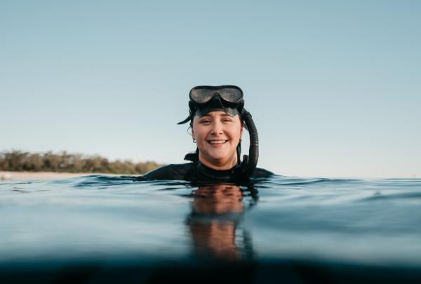 Amy Gash smiling in the ocean while wearing a snorkel with a tree-covered island in the distance, Lady Elliot Island, Southern Great Barrier Reef, Queensland © Tourism Australia