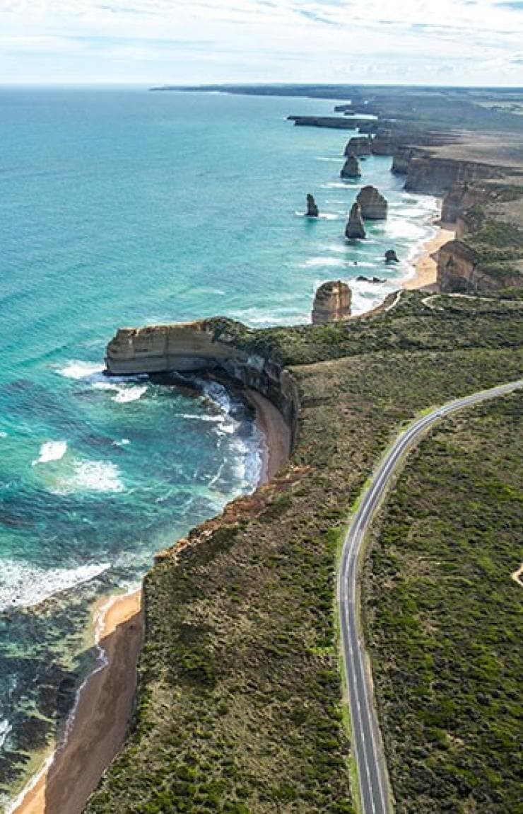 Aerial view over a road parallel to a dramatic coastline with free-standing sandstone pillars in the ocean, Twelve Apostles, Great Ocean Road, VIC © Greg Snell, Tourism Australia