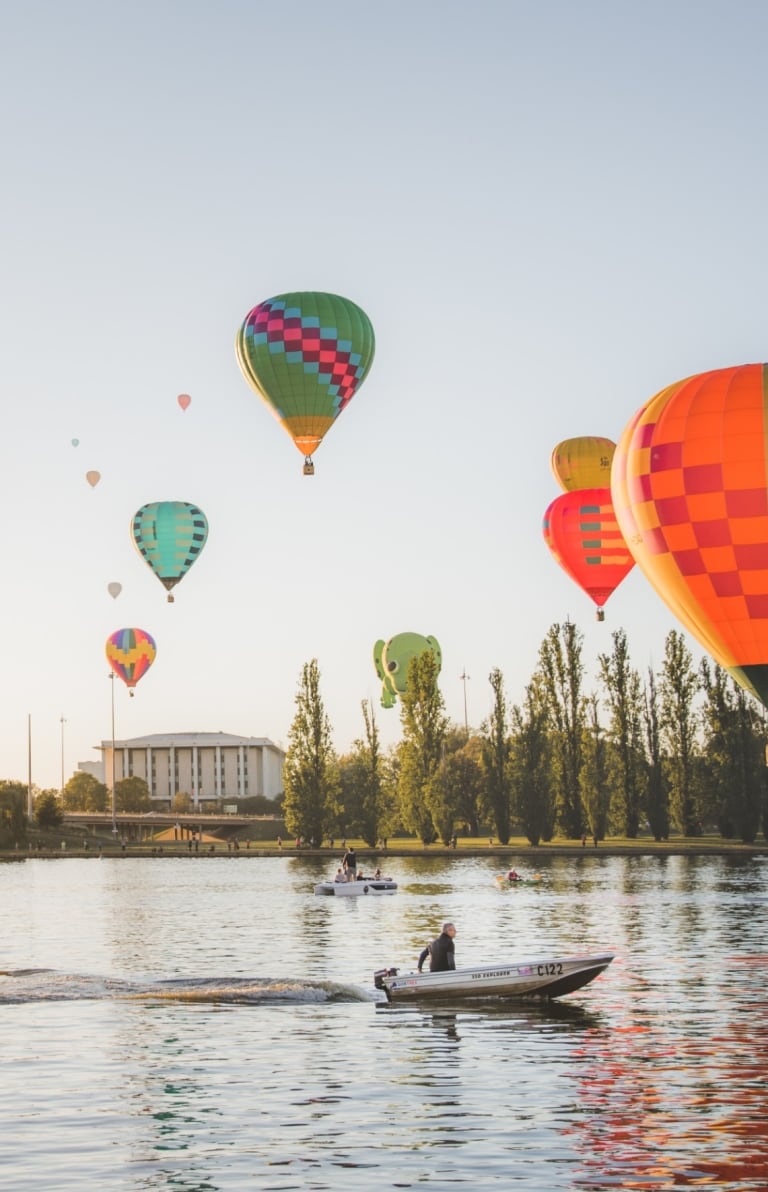 Montgolfières survolant les eaux paisibles du Lake Burley Griffin sur lequel voguent des bateaux à Canberra, Territoire de la Capitale Australienne © EventsACT