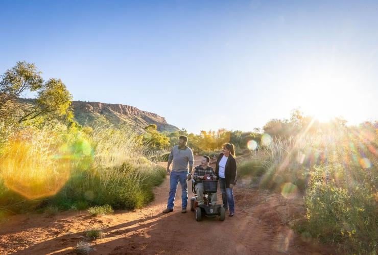 Homme en situation de handicap neurologique avec un scooter de mobilité en compagnie d'un autre homme et d'une femme dans l'Alice Springs Desert Park, Alice Springs, Territoire du Nord © Tourism NT/Helen Orr