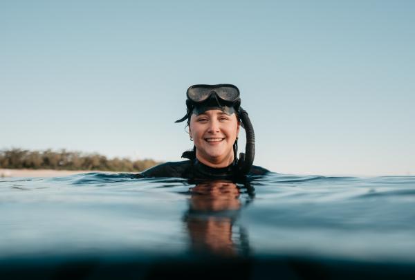 Amy Gash souriant dans l'océan tout en portant un masque et un tuba avec une île couverte d'arbres au loin, Lady Elliot Island, sud de la Grande Barrière de Corail, Queensland © Tourism Australia