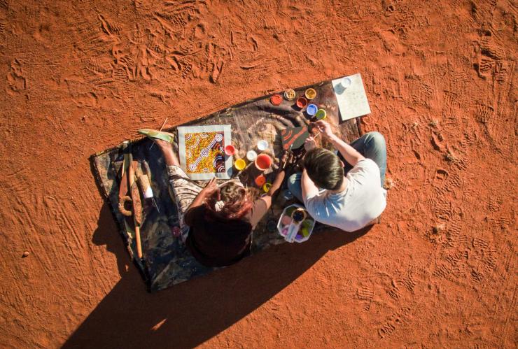Vue du dessus d'un visiteur apprenant la peinture à points avec un artiste aborigène local de Maruku Arts, près d'Uluru, dans le Territoire du Nord © Tourism NT/Archie Sartracom