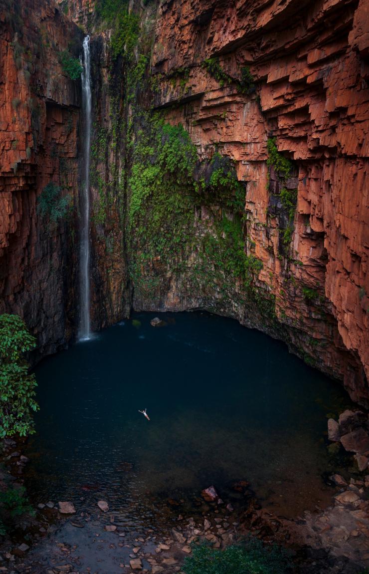 Vue aérienne d'une personne flottant sur le dos dans un point d'eau bleue entouré d'immenses parois rocheuses rouges ornées de mousse et d'une petite cascade à Emma Gorge, El Questro Wilderness Park, Kimberley, Australie Occidentale © Tourism Australia