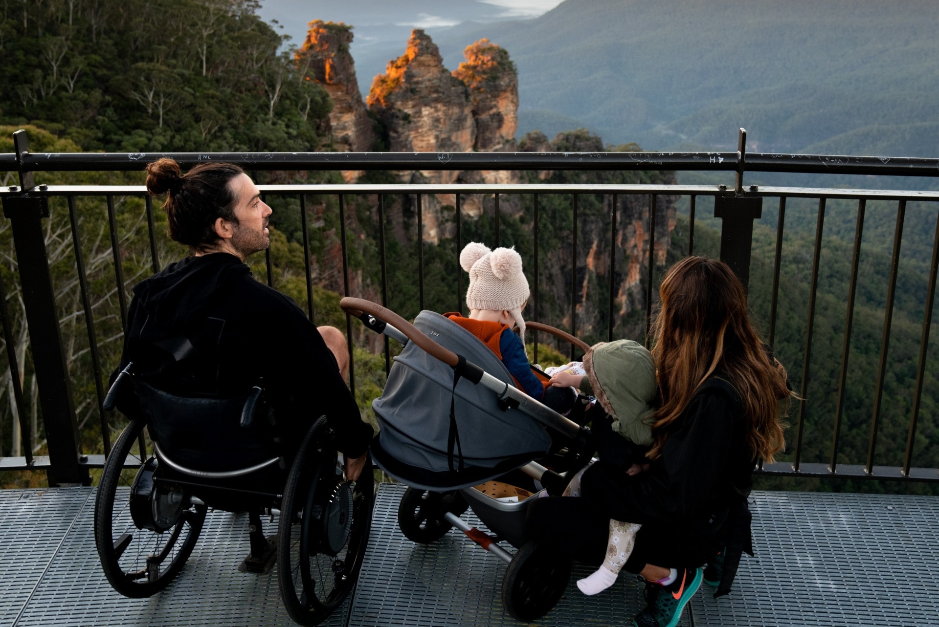 Seorang pria duduk di kursi roda, bersama keluarganya menatap Three Sisters di Blue Mountains, New South Wales © Tourism Australia