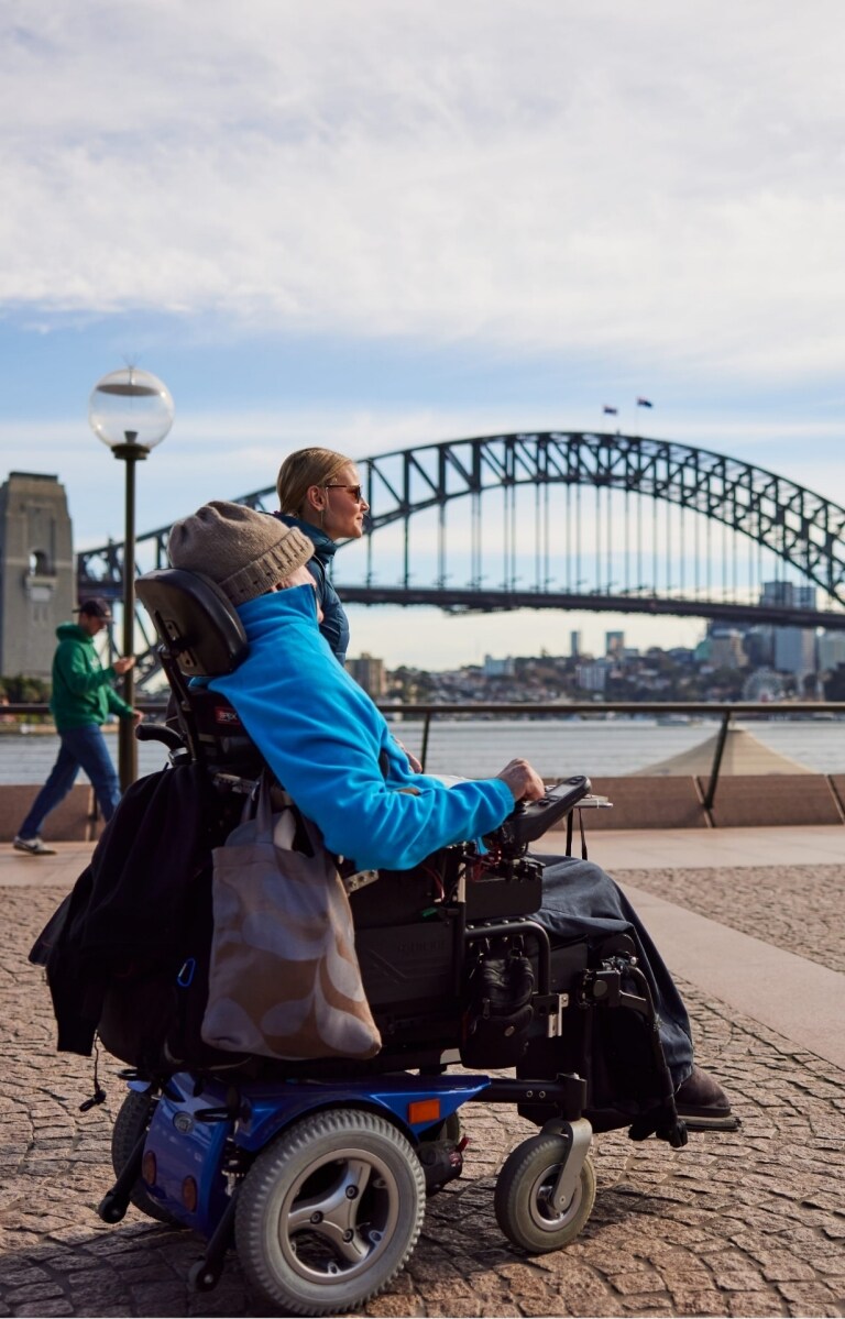 Orang yang menggunakan alat bantu mobilitas bersama orang lain yang berjalan di sampingnya mendekati Sydney Opera House dengan Sydney Harbour Bridge di latar belakangnya, di Sydney, New South Wales © Destination NSW