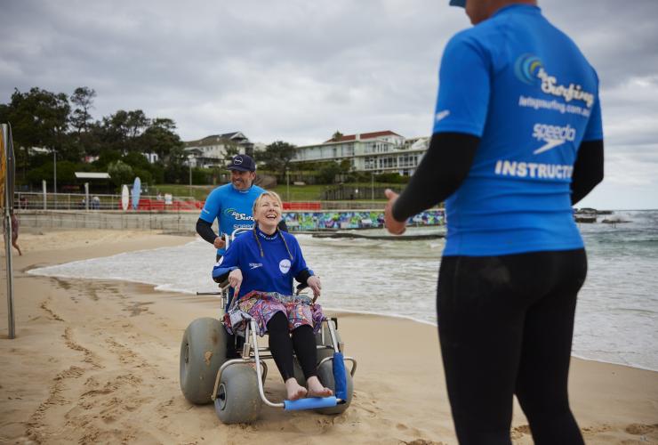 Seorang wanita duduk di kursi roda pantai di pasir Bondi Beach dengan instruktur Let's Go Surfing, Sydney, NSW © Tourism Australia