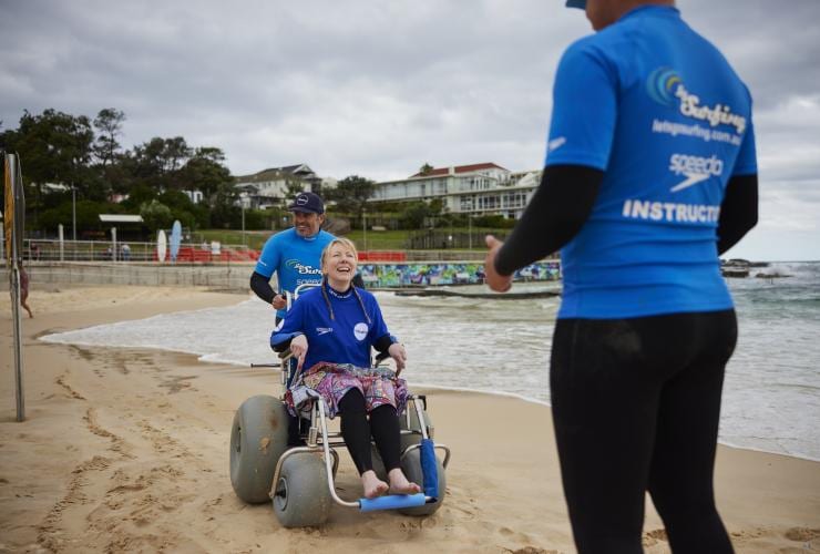 Seorang wanita duduk di kursi roda pantai di pasir bersama Let's Go Surfing, Bondi Beach, Sydney, New South Wales © Tourism Australia