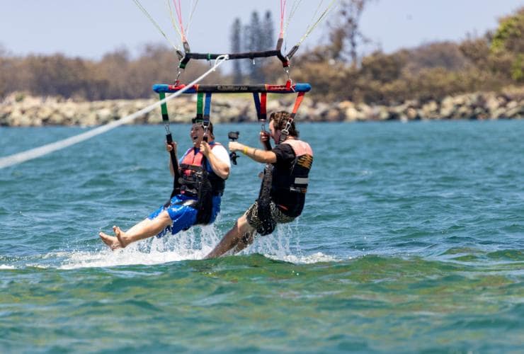 Seorang pria dengan neurodivergensi bermain paralayar dengan pria lain di Gold Coast Watersports, Surfers Paradise, Gold Coast, Queensland © Tourism and Events Queensland
