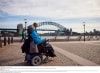 Orang yang menggunakan alat bantu mobilitas bersama orang lain yang berjalan di sampingnya mendekati Sydney Opera House dengan Sydney Harbour Bridge di latar belakangnya, di Sydney, New South Wales © Destination NSW