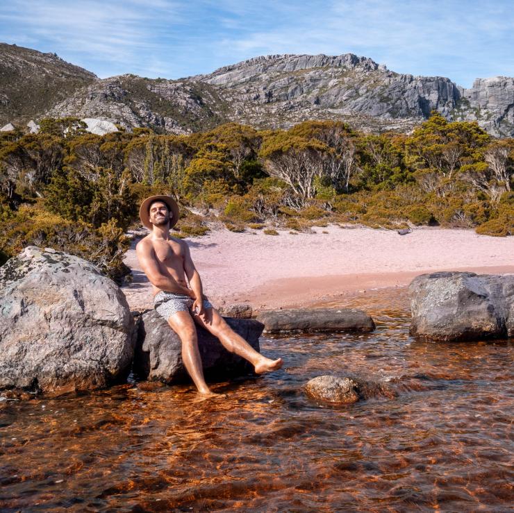 Miles Gray berpose di atas batu di sebuah danau di West-Coast Tasmania © Miles Gray, @milesuncharted
