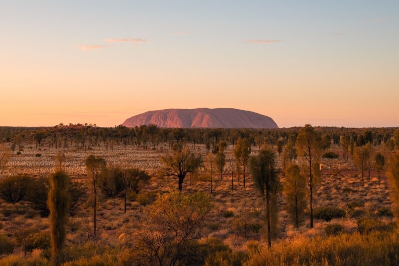 Un grande monolite di roccia rossa emerge dal paesaggio piatto dell'outback a Uluru, Uluru-Kata Tjuta National Park, Northern Territory © Tourism Australia