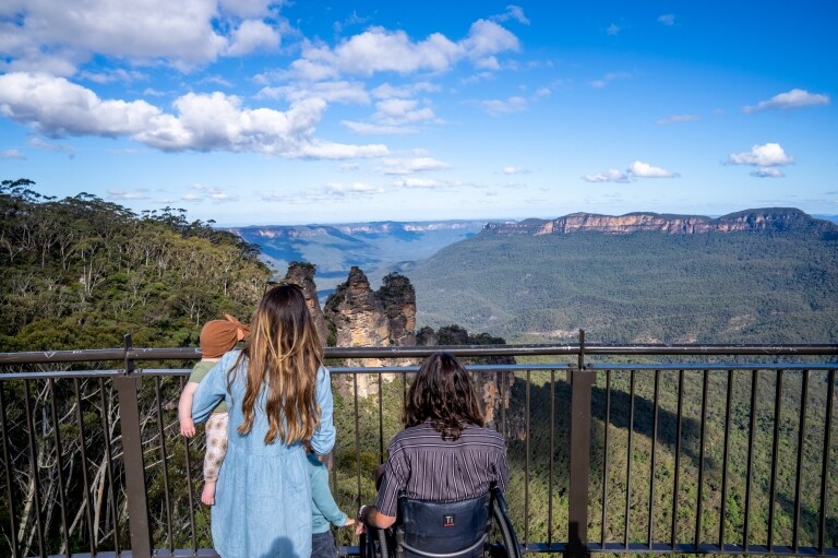 Uomo su una sedia a rotelle con la sua famiglia che ammirano le Three Sisters nel Blue Mountains National Park, New South Wales © Tourism Australia