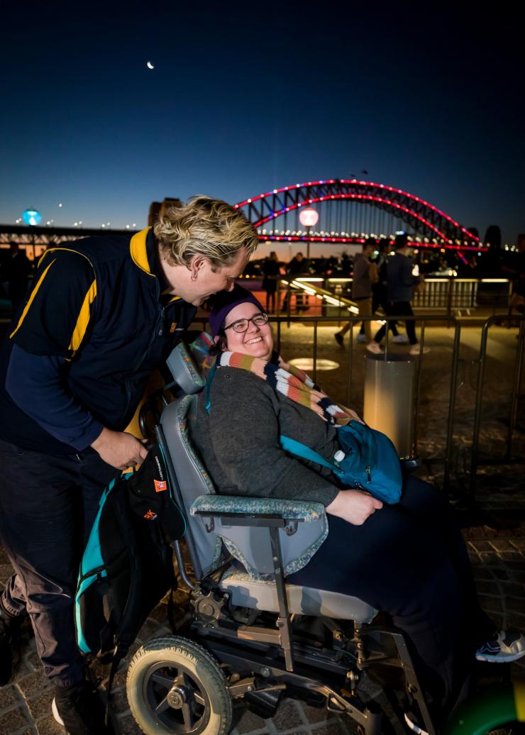 Donna su una sedia a rotelle con un'altra persona dietro di lei e il Sydney Harbour Bridge sullo sfondo, Vivid, Sydney, New South Wales © Destination NSW