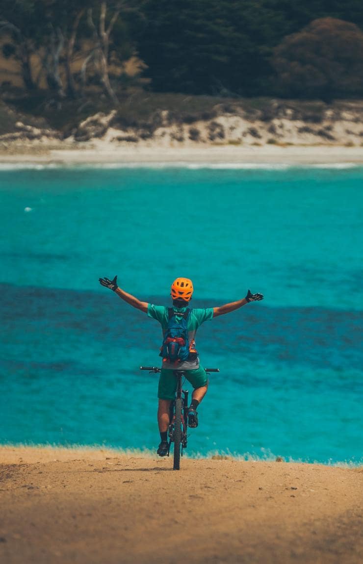 Persona in mountain bike con le braccia alzate affacciata sull'oceano blu nel Maria Island National Park, Tasmania © Matt Staggs
