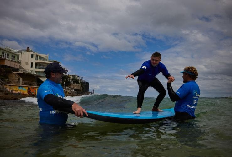 ニュー・サウス・ウェールズ州、シドニー、ボンダイ・ビーチ、レッツゴー・サーフィンの補助でサーフボードの上に立つ神経的多様性を持つ男性 © Tourism Australia