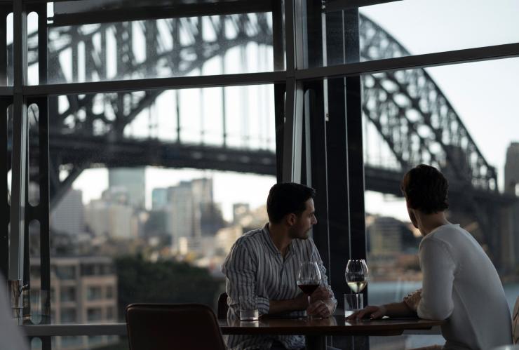 キー・レストランからシドニー・ハーバー・ブリッジ越しにシドニー・ハーバーの景色を眺めながらワインを楽しむカップル © Destination NSW