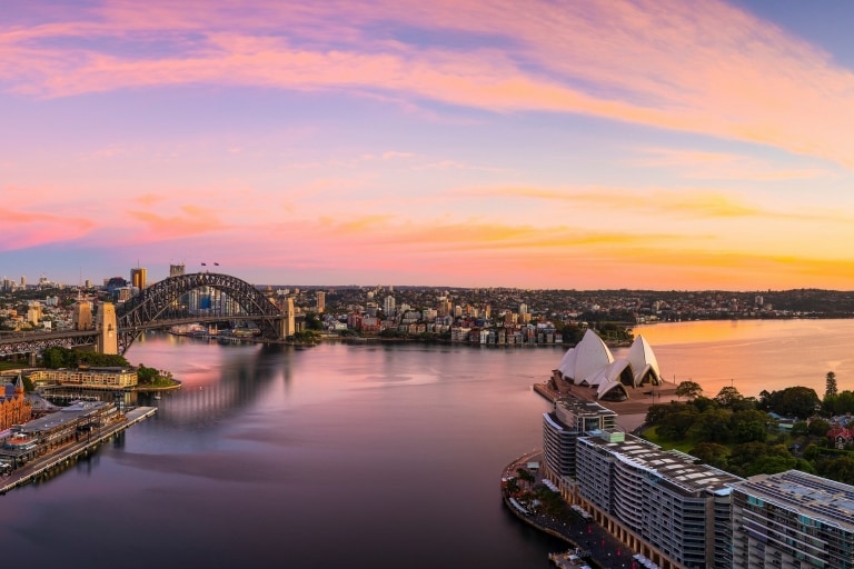 ニュー・サウス・ウェールズ州、シドニーの夕日の青、ピンク、金色の色彩を映したシドニー・ハーバーの上空からの眺め © Destination NSW