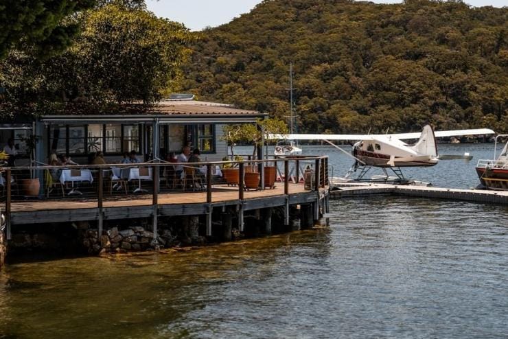 부두 옆에 수상비행기가 정박해 있는 가운데, 숲지대로 둘러싸인 강변 레스토랑에서 식사를 하는 사람들