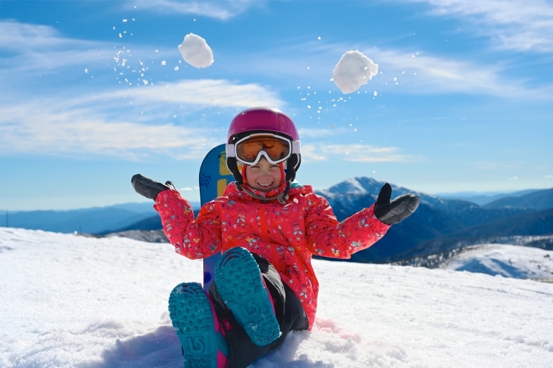 Một em bé ngồi trên đỉnh núi phủ tuyết trắng, tung những quả cầu tuyết lên không trung, Hotham Alpine Resort, Núi Hotham, Victoria © Hotham Alpine Resort