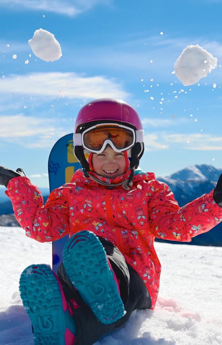 Một em bé ngồi trên đỉnh núi phủ tuyết trắng, tung những quả cầu tuyết lên không trung, Hotham Alpine Resort, Núi Hotham, Victoria © Hotham Alpine Resort