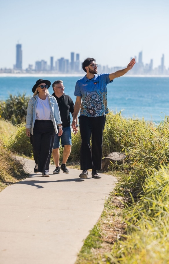 A friendly guide from the Jellurgal Aboriginal Cultural Centre engages warmly with a group of tour participants. Surrounded by the lush, green beauty of Burleigh Heads on the Gold Coast, and the sparkling ocean under a bright blue sky, the scene highlights the connection to the land, Burleigh Heads, Queensland © Tourism Australia / Jellurgal Aboriginal Cultural Centre