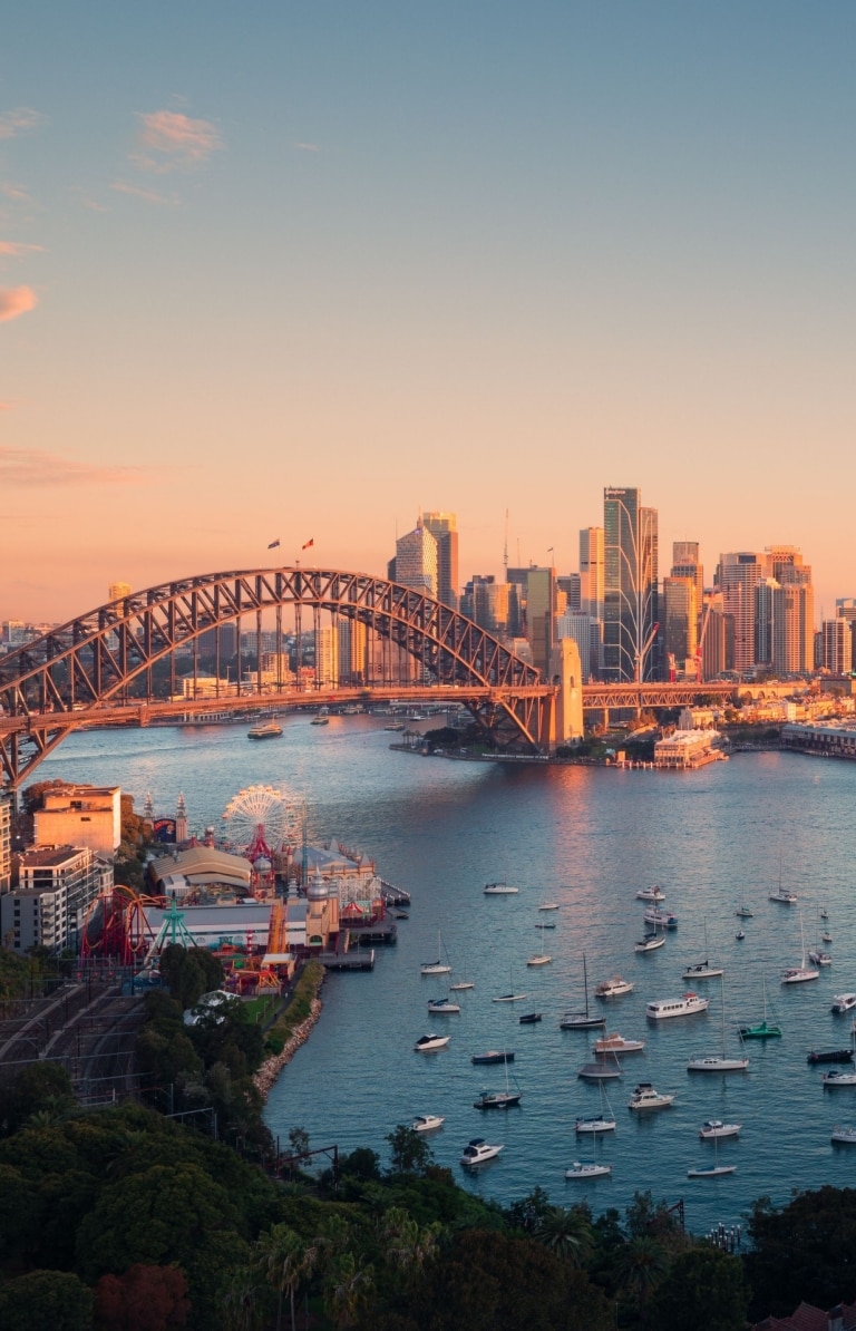 Aerial view over a tranquil blue harbour sprinkled with boats with an arched bridge leading to a city skyline in Sydney Harbour, Sydney, New South Wales ©Tourism Australia