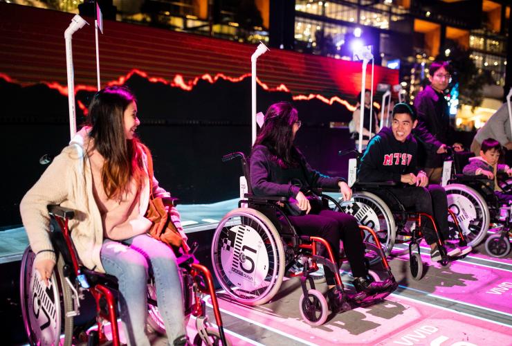 A group of friends seated in wheelchairs in a reserved area, with colourful lights surrounding them at Vivid Sydney, New South Wales © Destination New South Wales