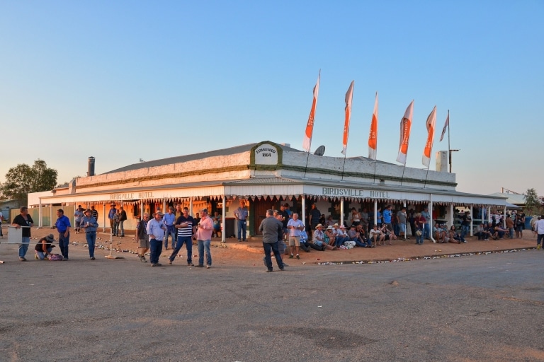 A historic pub with a wrap-around awning decorated with flags and a crowd of people gathered around outside sitting, drinking and talking at Birdsville Hotel, Birdsville, Queensland © Tourism and Events Queensland