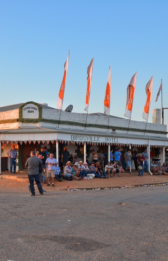 A historic pub with a wrap-around awning decorated with flags and a crowd of people gathered around outside sitting, drinking and talking at Birdsville Hotel, Birdsville, Queensland © Tourism and Events Queensland