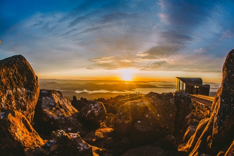Large rocks in the foreground with two people standing at a lookout in the distance admiring views over Hobart as the sun beams over the landscape at kunanyi/Mt Wellington, Hobart, Tasmania © Tourism Australia