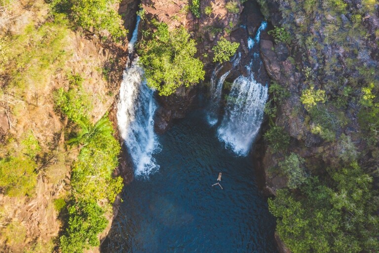 Aerial view over a person floating in a natural blue pool surrounded by trees with two large waterfalls cascading down at Florence Falls, Litchfield National Park, Northern Territory © Tourism NT/Dan Moore