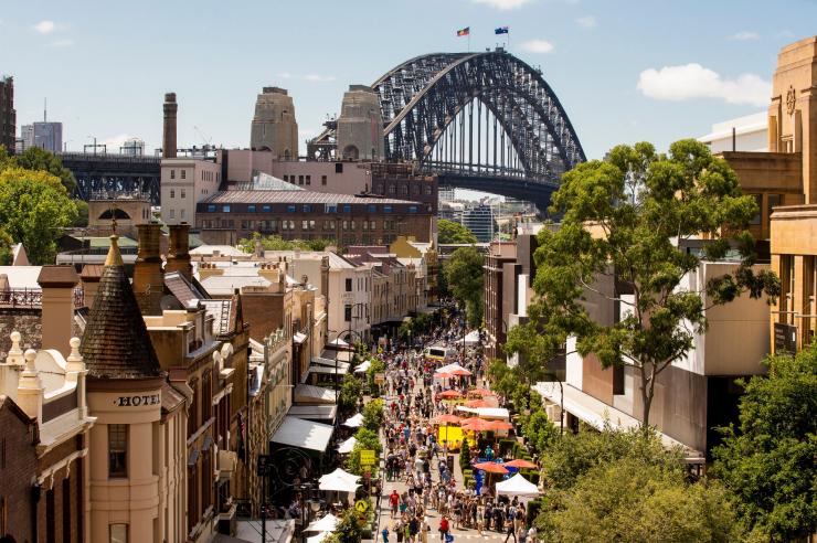 新南威尔士州,明媚阳光下,人们漫步在花伞林立的街道上的鸟瞰图,背景是悉尼海港大桥 © 新南威尔士州旅游局版权所有 新南威尔士州,明媚阳光下,人们漫步在花伞林立的街道上的鸟瞰图,背景是悉尼海港大桥 © 新南威尔士州旅游局版权所有