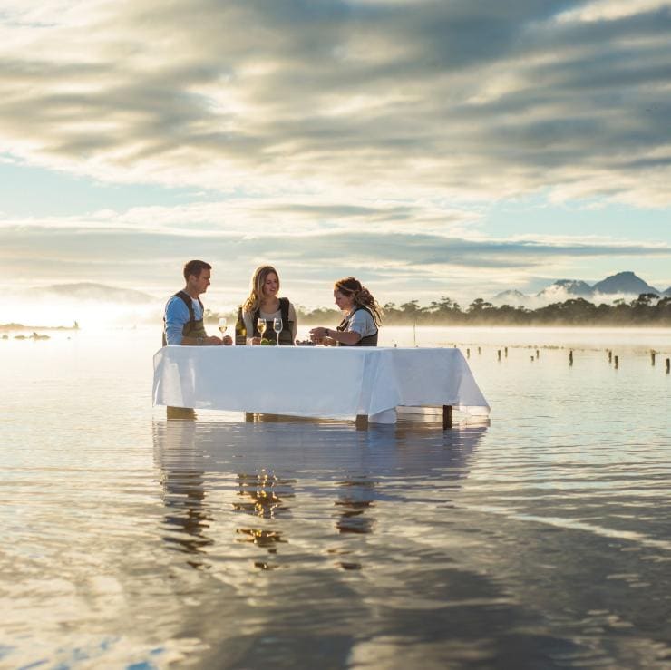三個人站在塔斯曼尼亞歌斯灣薩菲爾菲欣納海洋生蠔養殖場的水中餐桌旁吃生蠔©薩菲爾菲欣納/塔斯曼尼亞旅遊局