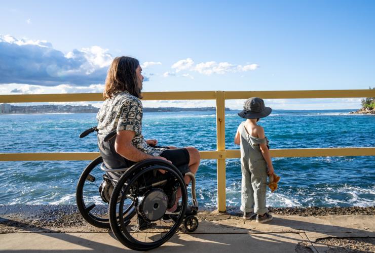 Ein Mann in einem Rollstuhl blickt mit seinem Sohn in Manly aufs Meer, Sydney, New South Wales © Tourism Australia