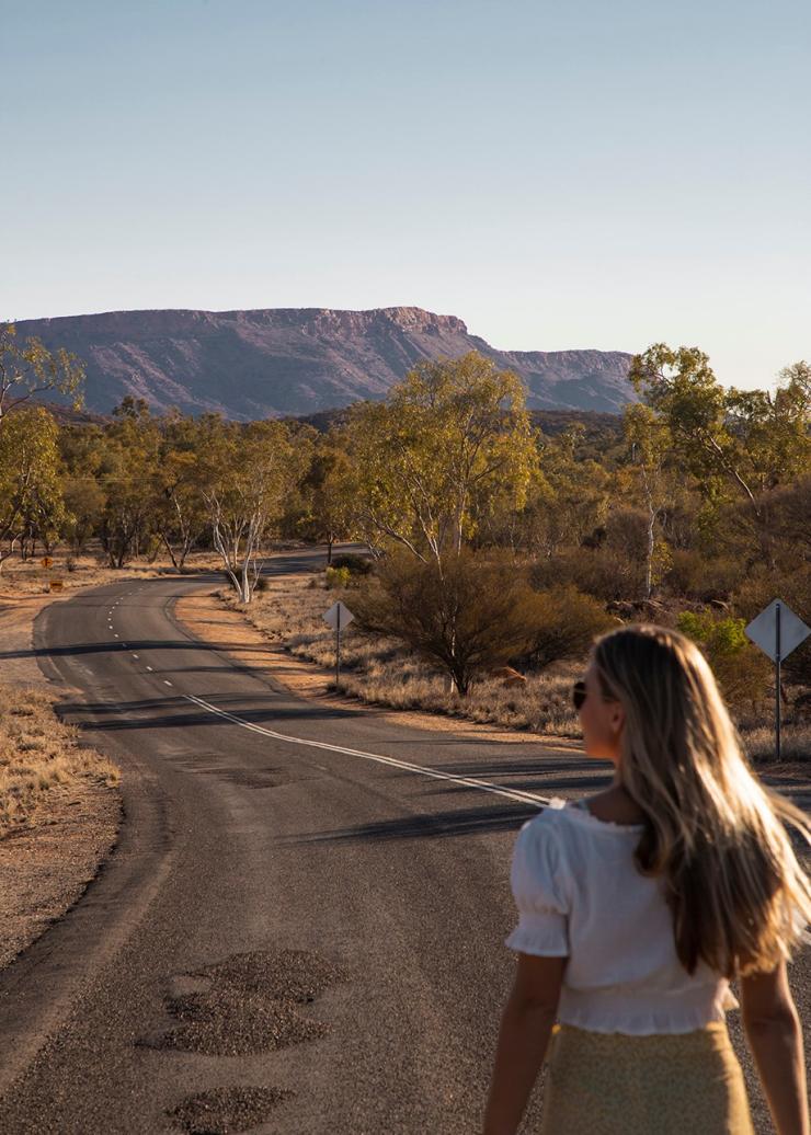 Eine Person steht auf einer Straße, die sich durch Buschland zu einer großen Felsformation in der Nähe der Alice Springs Telegraph Station windet, Alice Springs, Northern Territory © Tourism Australia