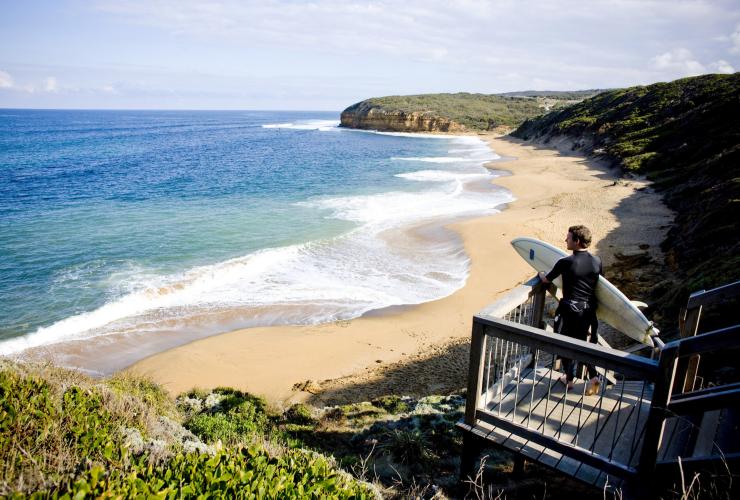 Ein Surfer steht mit einem Surfbrett unter dem Arm an einem goldenen Strand am Great Ocean Road, Victoria © Tourism Australia