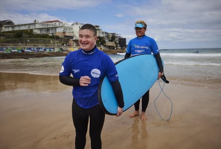 Ein junger Mann mit einer geistigen Behinderung trägt lächelnd mit der Hilfe eines Surflehrers ein Surfboard am Bondi Beach, Sydney, New South Wales © Tourism Australia