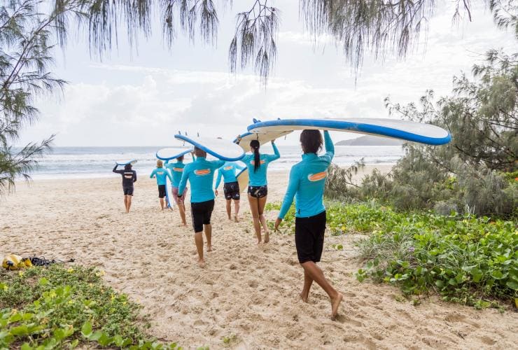 Eine Gruppe von Menschen mit Surfbrettern auf den Köpfen, während sie auf goldenem Sand in Richtung Meer laufen, am Noosa Main Beach, Noosa, Queensland © Tourism and Events Queensland